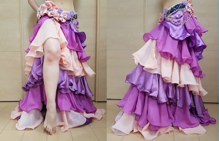 the image shows two angles of a belly dance costume. the costume features a layered skirt made of purple, pink, and white chiffon and satin fabrics, arranged in ruffles. the waist belt is adorned with floral embellishments. a person is wearing the costume and is standing barefoot in both images.