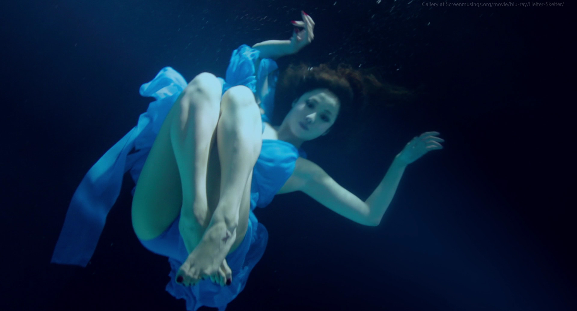 a young woman in a flowing blue dress is suspended underwater. she appears graceful and elegant as she floats with her arms outstretched and her hair swirling around her. the lighting is artificial, creating a dreamlike atmosphere. the overall color palette is predominantly blue, ranging from deep navy to a brighter turquoise.