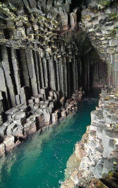 a stunning shot inside fingal's cave, showcasing its iconic basalt columns that form the walls and ceiling of the cave. a body of water fills the cave floor, reflecting the dark, textured rock formations above. the water has a greenish tint, and the lighting inside the cave is subdued, creating a mysterious and aweinspiring atmosphere.