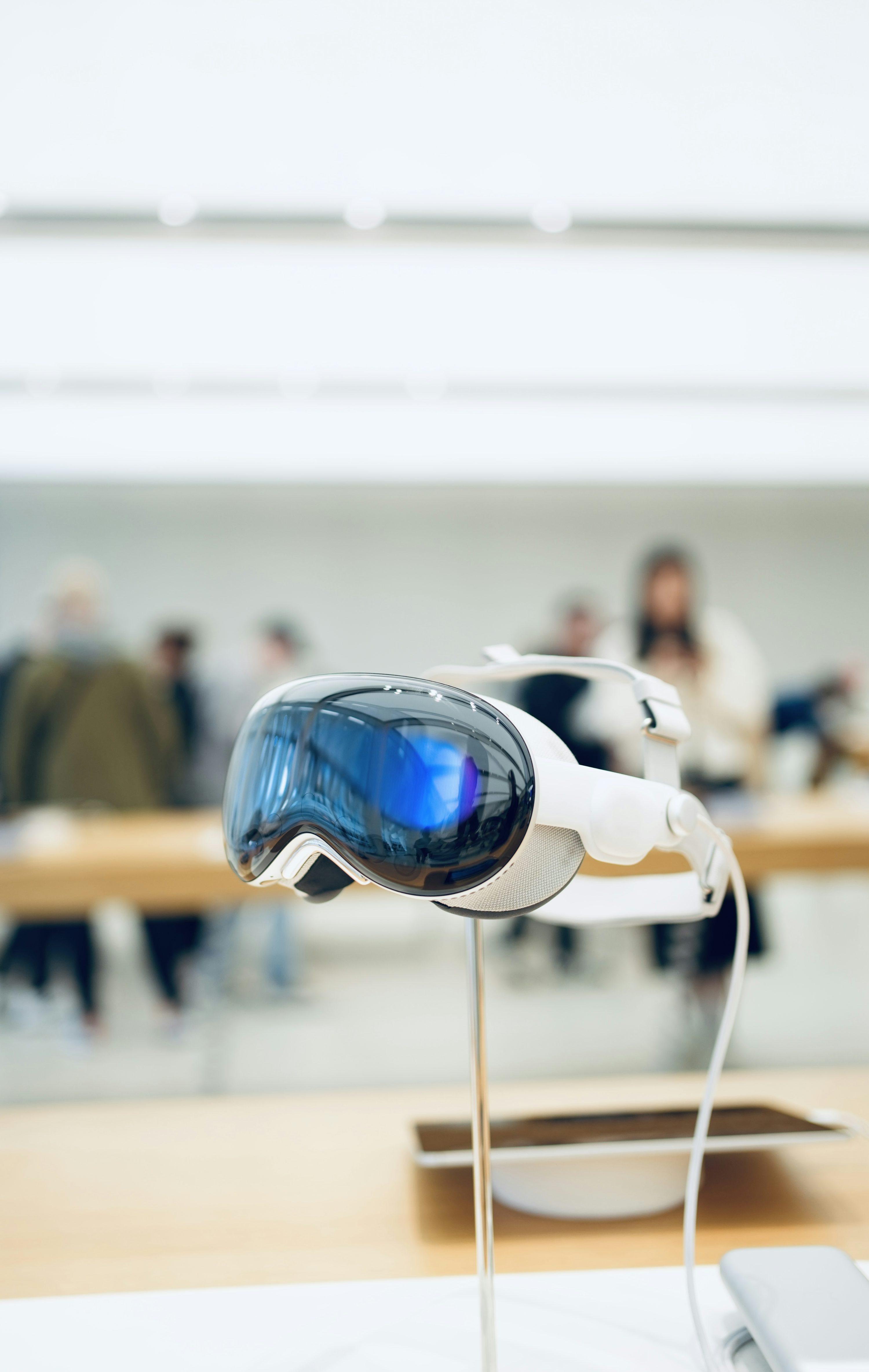 a closeup shot shows an apple vision pro vr headset displayed on a thin, silver stand atop a light wooden table. the lenses of the headset feature a glossy, reflective surface with a vibrant blue hue, while the headset itself is sleek white. a thin white cable is connected to the side. the background is an outoffocus retail environment, hinting at a technology store with people browsing in the background and a bright, evenly lit setting.