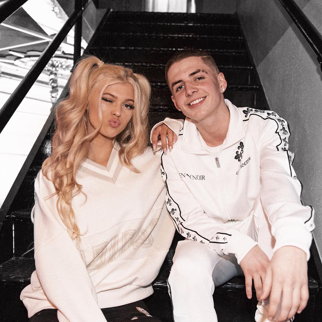 two people, a man and a woman, are sitting on a set of stairs. the woman on the left has blonde hair in pigtails and is winking with a pout. she is wearing a white sweatshirt. the man on the right has short brown hair and is smiling. he is wearing a white tracksuit. they are both sitting on black stairs with black railing.