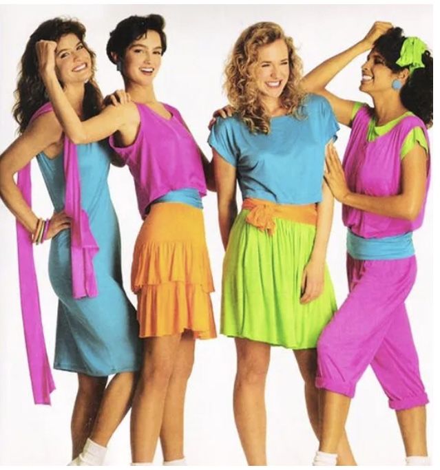 four women are posing for a photo, wearing bright, colorful outfits reminiscent of 1980s fashion. from left to right: the first woman is wearing a blue dress with a pink scarf. the second woman is wearing a pink top and orange skirt. the third woman is wearing a blue top and green skirt. the fourth woman is wearing a pink top and pants with blue accents, and a green headband. they are all smiling and appear to be enjoying themselves. the background is plain white.