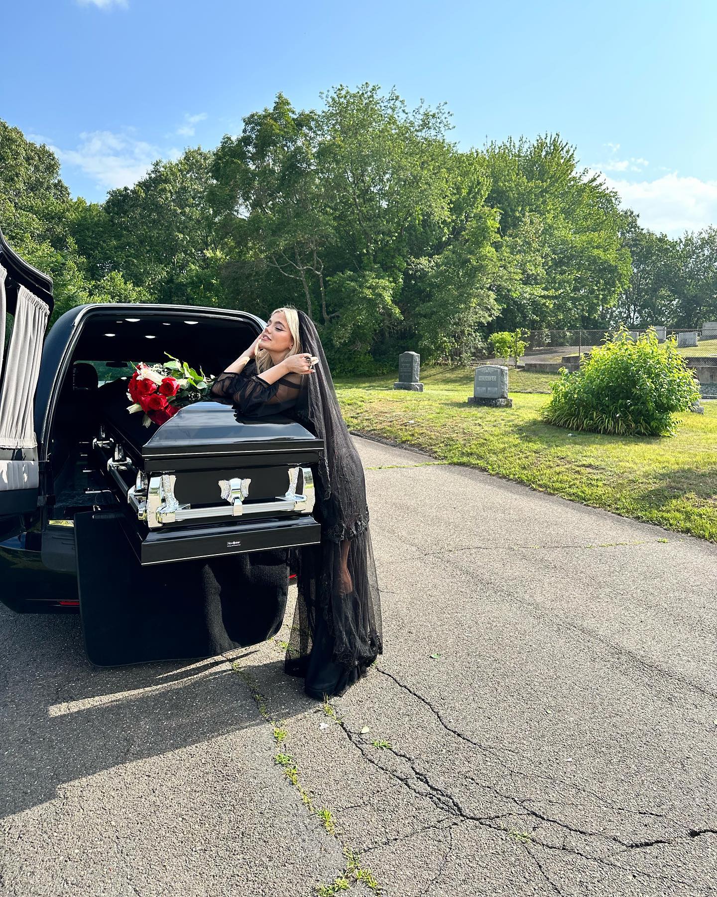 a woman wearing a black dress and veil is leaning on a black coffin with roses inside a hearse in a cemetery. trees and gravestones are visible in the background.