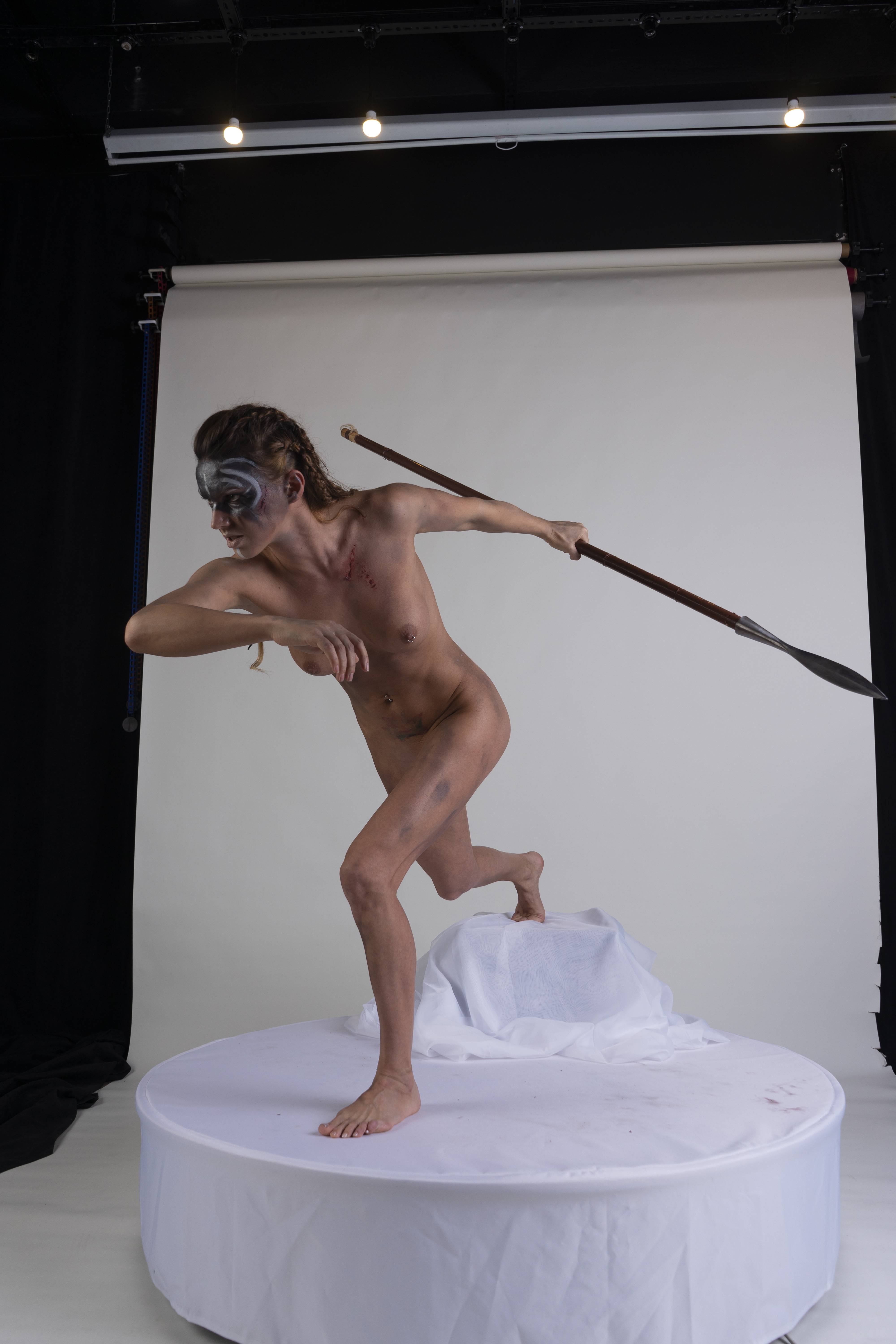 a nude woman with fair skin and an athletic build is holding a spear and posing in a dynamic action pose. she has war paint on her face and braids in her hair. her body has bruises, blood, and a wound, suggesting she's a warrior or a mythological figure in the midst of battle. the photo is a studio shot with a white background and a round platform. the pose and expression convey determination, strength, bravery, courage, and aggression.