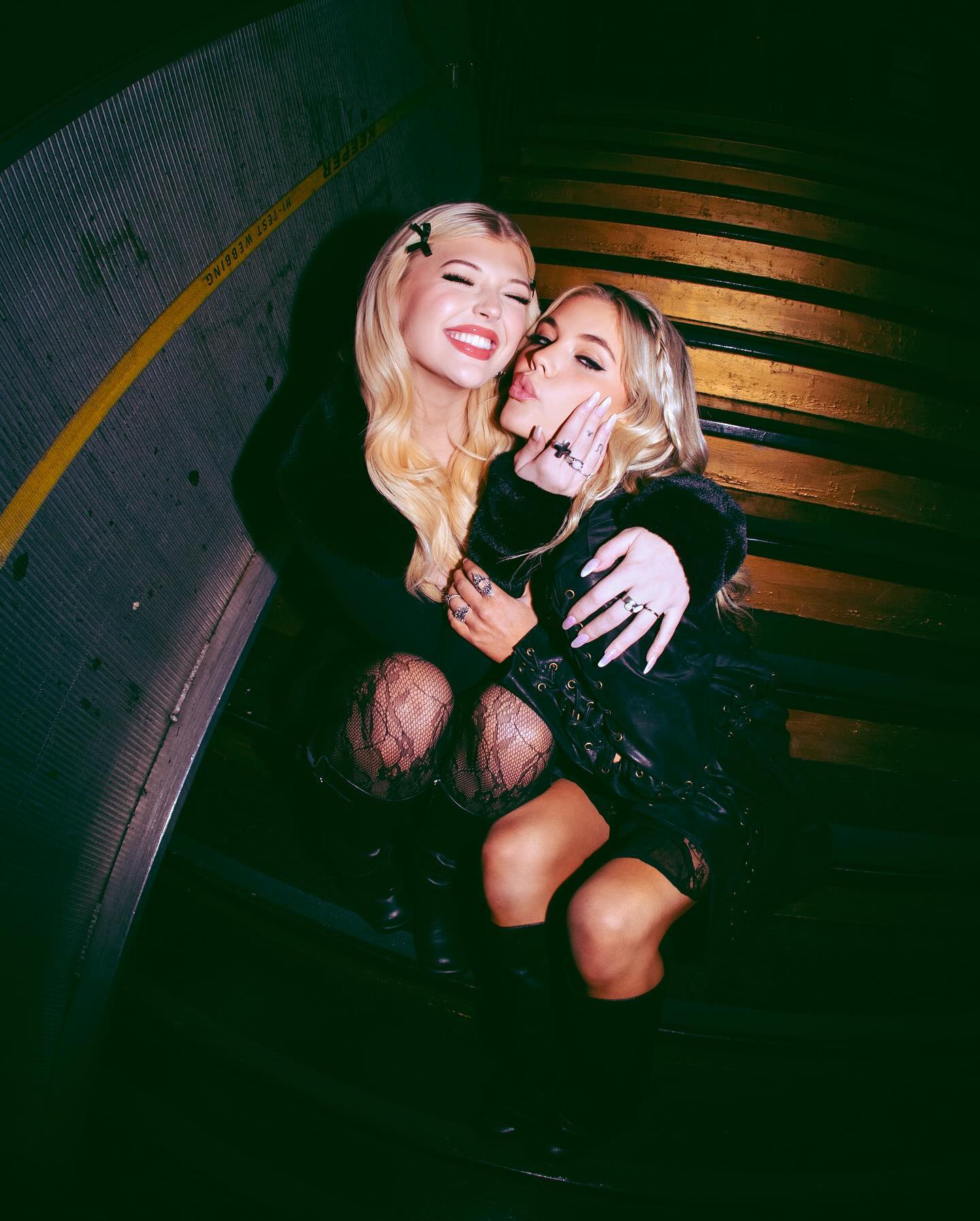 two young women are posing for a photo indoors, likely on a staircase. the woman on the left has long blonde hair with a hair clip, wearing a black dress, fishnet stockings, and black boots, and is smiling widely. the woman on the right has blonde braided hair, is wearing a black jacket and dress, and has her hand on her cheek while pouting her lips. both are wearing jewelry, specifically rings on their fingers. the lighting is dark, creating a moody atmosphere.