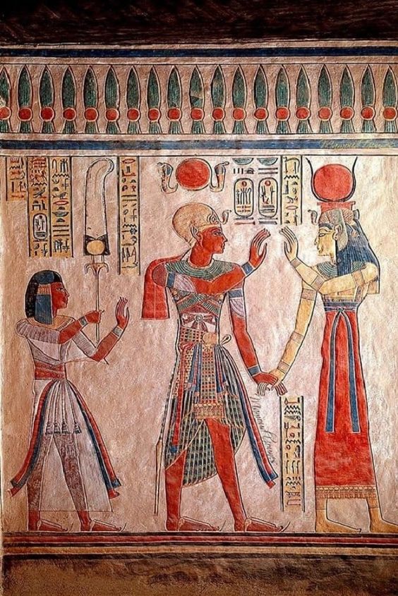 a painting showing three people, two men and a woman, in ancient egyptian attire. the men wear skirts and decorative collars. the woman wears a long dress. the man in the middle is gesturing toward the man on the right, who is wearing a headdress.  all three figures have dark hair and brown skin. there are hieroglyphs surrounding the figures and a decorative border along the top of the image.