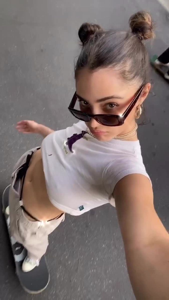 a young woman with brown hair styled in two buns is skateboarding while looking up at the camera. she's wearing sunglasses, a white cropped tshirt, and grey pants. she has a gold chain necklace and hoop earrings. another person's skateboard is visible in the background.