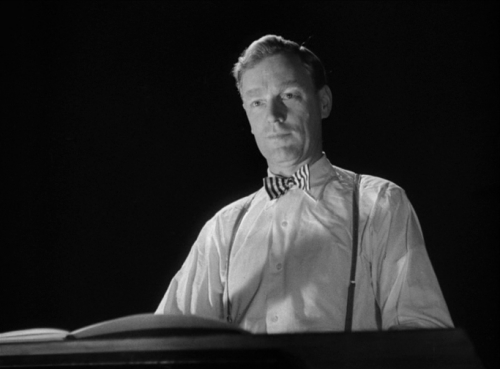 a man wearing a white shirt, bowtie and suspenders sits at a table, his face illuminated by a light, reading a book. he has a thoughtful expression on his face. the photo is in black and white, and the background is dark.