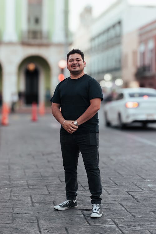 a fullbody shot of a smiling middleaged hispanic man standing on a city street. he's wearing a black tshirt, black pants, and black converse sneakers.  the background is slightly out of focus, showcasing city buildings and a car. the overall impression is one of casual confidence and positivity.