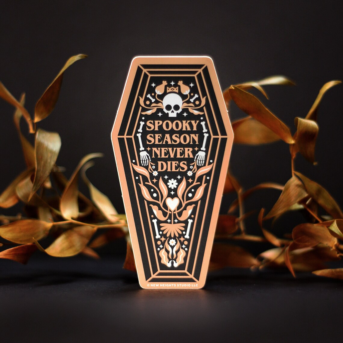 the image showcases a stylish, diecut sticker shaped like a coffin.  the sticker is predominantly black with a rose gold/peach colored design. the design features a skull, crossbones, floral elements, and the text "spooky season never dies."  the sticker is positioned against a backdrop of dried, goldenbrown autumn leaves, enhancing the halloween and autumnal theme. the overall style is a blend of vintage/retro aesthetics with a touch of dark academia and gothic elements.  a small copyright notice for "new heights studio llc" is visible at the bottom. the image is welllit and professionally photographed, highlighting the details of the sticker's design and the autumnal setting.