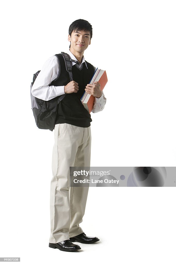 a young asian male stands against a white background. he is wearing a white collared shirt, a black vest, and tan pants, and black dress shoes. he is carrying a backpack and holding books in his right hand.