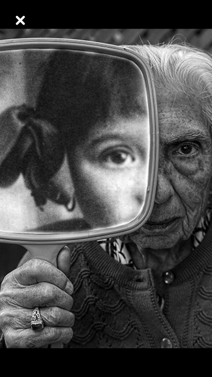 an elderly woman with grey hair holds a mirror up to her face, the reflection shows a younger version of herself.  she is looking down, and her face is full of wrinkles. she appears sad, pensive, and alone.  the image is in black and white, which adds to the somber mood.  the image suggests a sense of loss, regret, or sadness. the woman's hand is adorned with a ring.