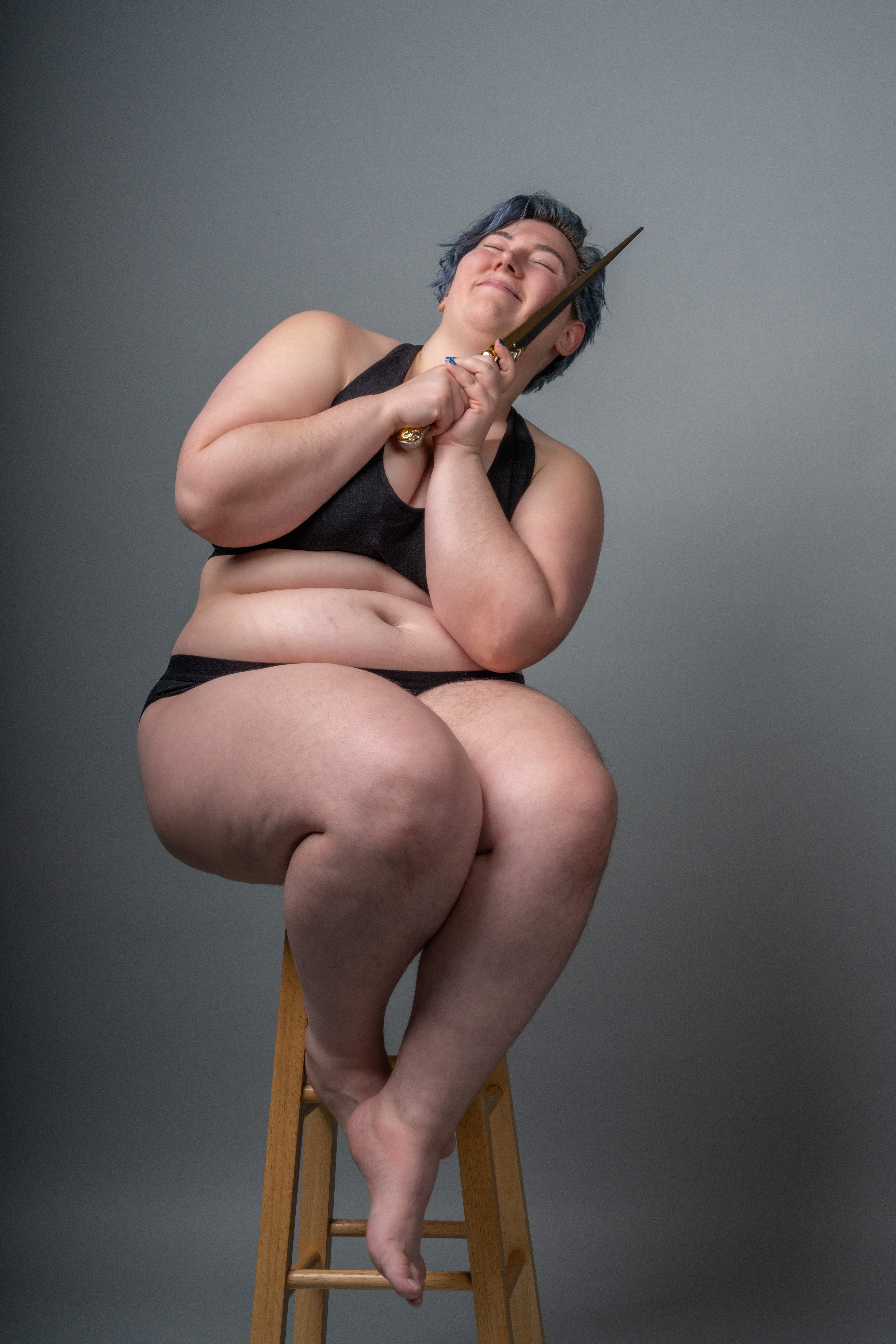 a plus size woman with blue hair, wearing black underwear and a bra, is sitting on a wooden stool in a studio with a gray background. she is holding a wand with both hands near her face, smiling with her eyes closed, and appearing confident. she is barefoot.