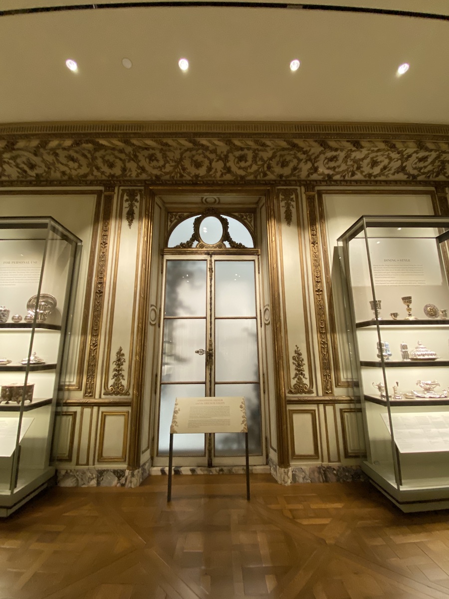 the photograph captures an interior museum setting with a focus on decorative arts. display cases containing silver artifacts are positioned on either side of a set of french doors. the doors are framed by ornate molding, and a sign stands in front. the walls are decorated with paneling and marble baseboards, and the floor is covered in parquet. lighting fixtures provide illumination from the ceiling.