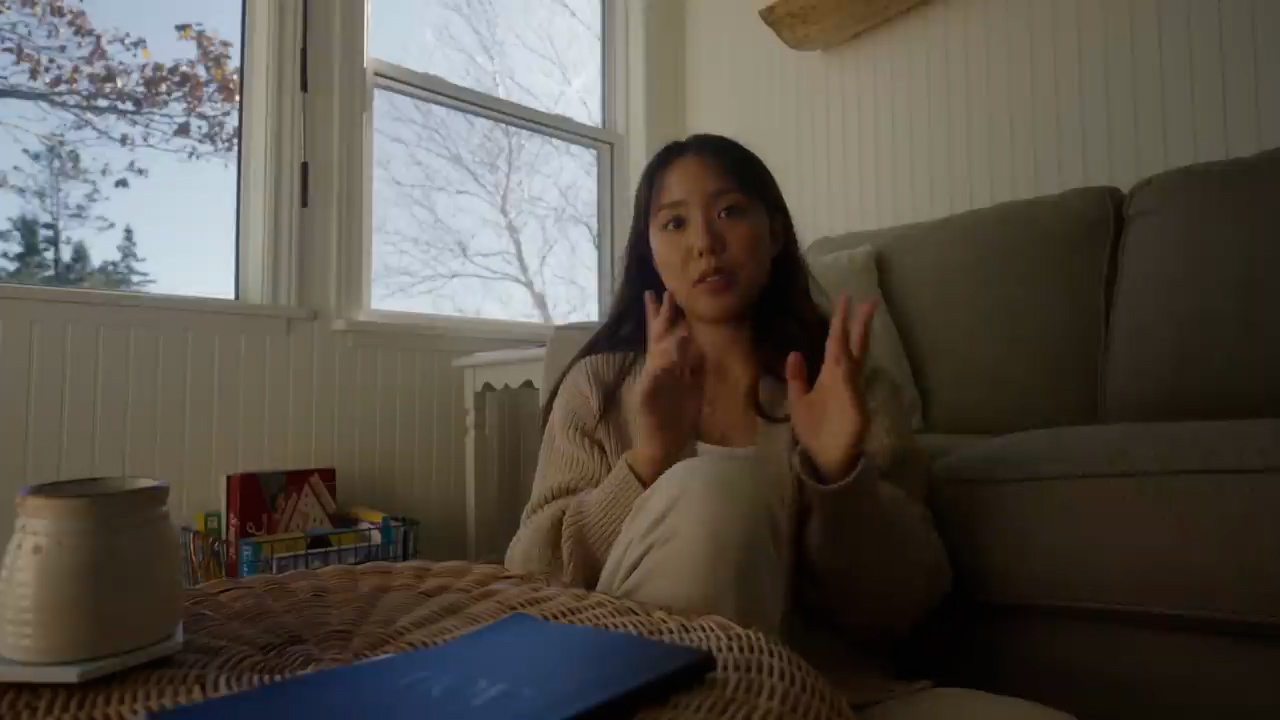 a young asian woman sits on the floor of her living room, talking to the camera. she is recording a vlog or video call. the room is cozy and filled with natural light, with a couch behind her and a window to the left. there are books and a coffee mug on a wicker table in front of her.