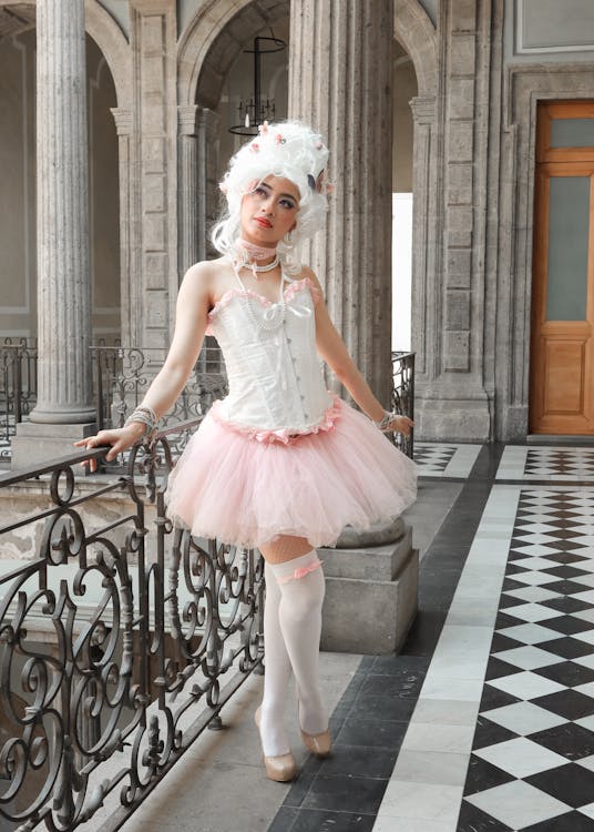 a young woman in a rococostyle cosplay costume stands on a balcony in a grand building. she wears a white corset, pink tutu, white stockings and heels, and a large white wig adorned with pink flowers. the building's interior features ornate architecture and black and white tiled floors.  the overall aesthetic is elegant and graceful.