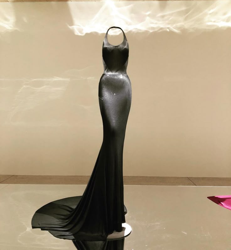 a black halter neck evening gown displayed on a mannequin is shown in a full shot. the dress is sleek and has a slight train. it is displayed on a reflective surface that shows its reflection. the background is a plain lightcolored wall with some light and shadow patterns.