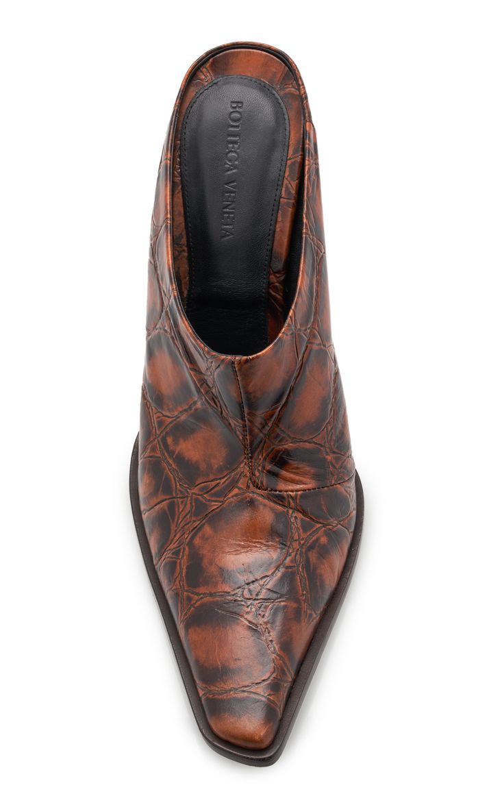 a studio shot against a white background shows a pair of bottega veneta women's brown leather mules with a pointed toe. the shoes are made of crocodileembossed leather and have a low heel. the insole is visible and has the bottega veneta logo printed on it.