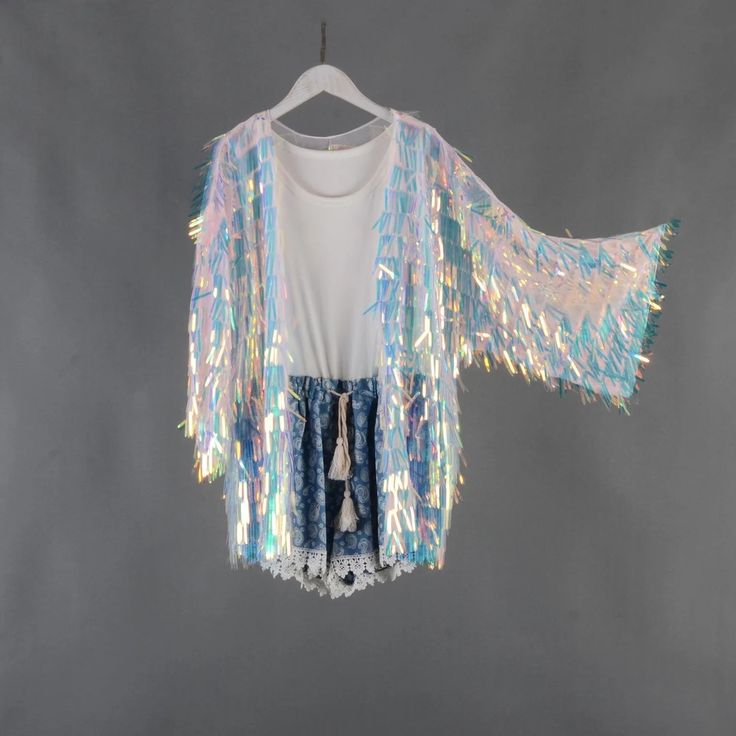 a white iridescent sequin kimono jacket with fringe hanging off the edges of the garment. it is hanging on a white hanger on a gray background. the jacket is open, revealing a white tank top and denim shorts with a lace trim.