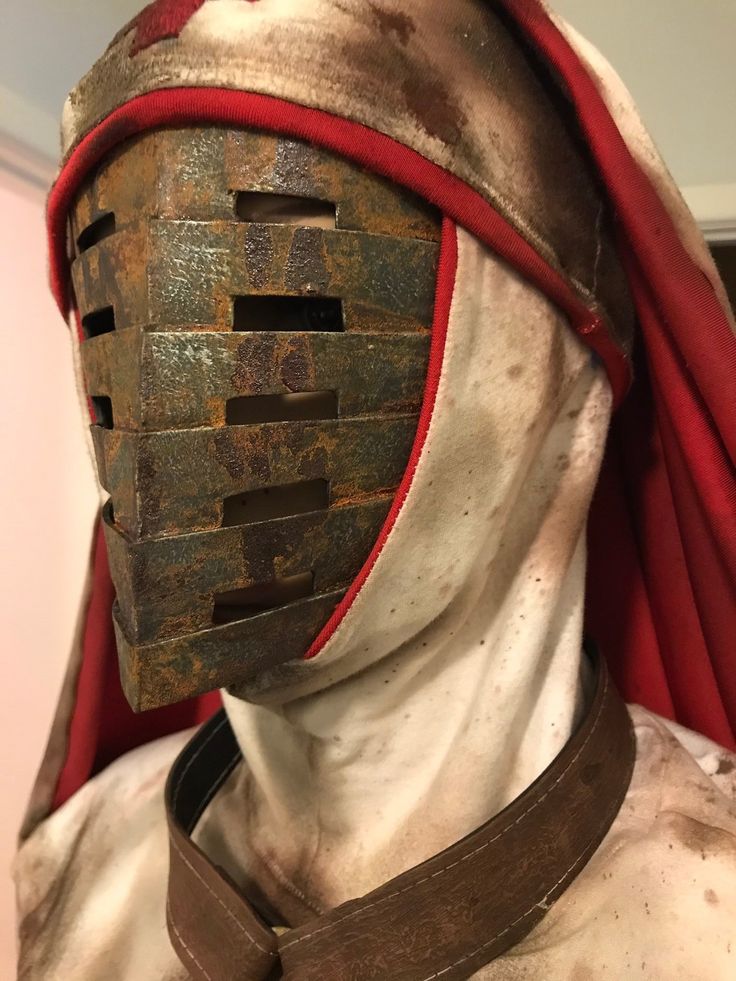 closeup of a person wearing a costume. the costume features a white, hooded head covering with red trim and a series of rusty metal plates forming a mask. the mask has horizontal slits for visibility. a brown leather neck brace is also part of the costume. the fabric and metal parts appear weathered and dirty.