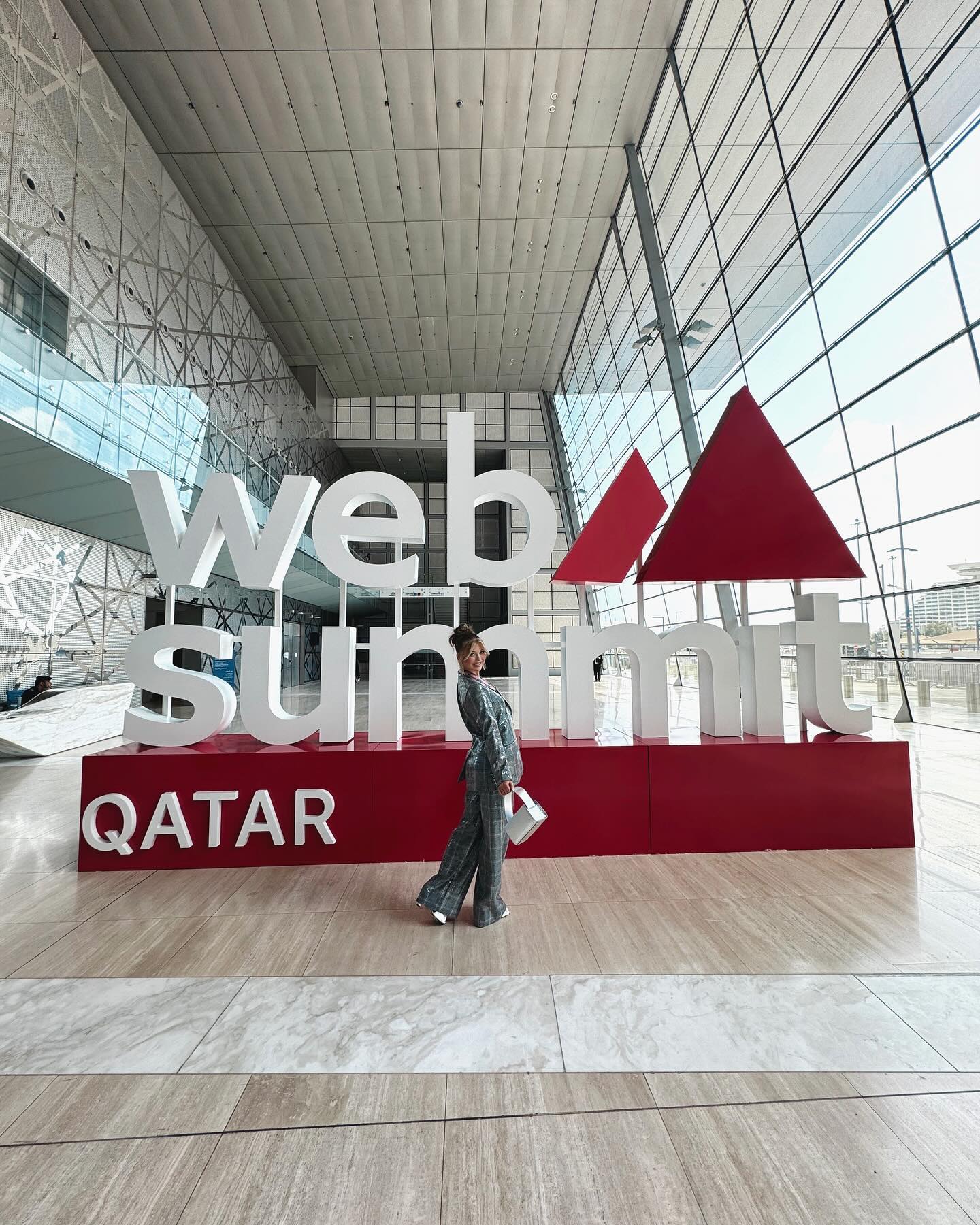 a woman stands in front of a large "web summit qatar" sign inside a modern convention center with a glass facade. the sign is white with red accents, and the building has a high ceiling and patterned flooring. the woman is wearing a patterned suit and holding a white bag.