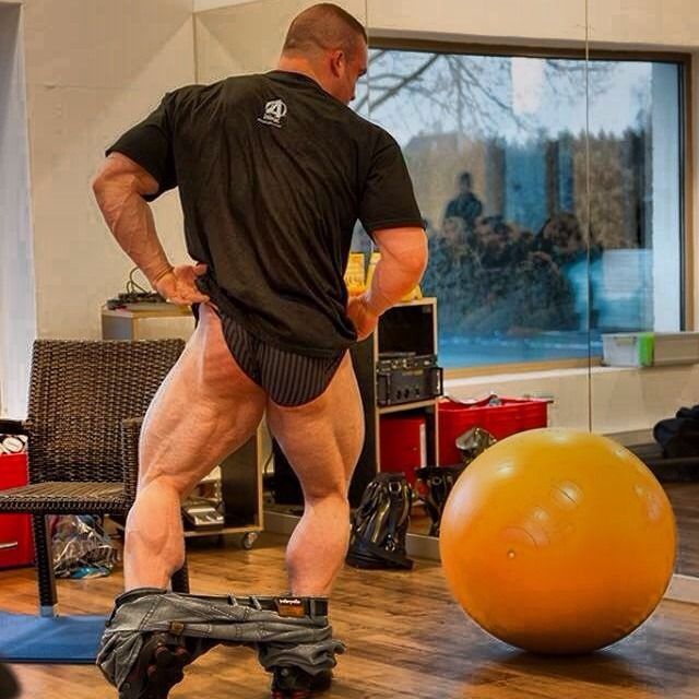 a muscular man in a black shirt and black shorts is standing in a gym, looking at a large orange exercise ball in the foreground. the man is clearly wellbuilt and is likely a bodybuilder. he appears to be in the middle of a workout, possibly taking a break or considering his next exercise. the gym has wooden floors, a large window, and various pieces of equipment in the background. the man's focus is on the exercise ball, and his determined expression suggests he is ready to continue his workout.