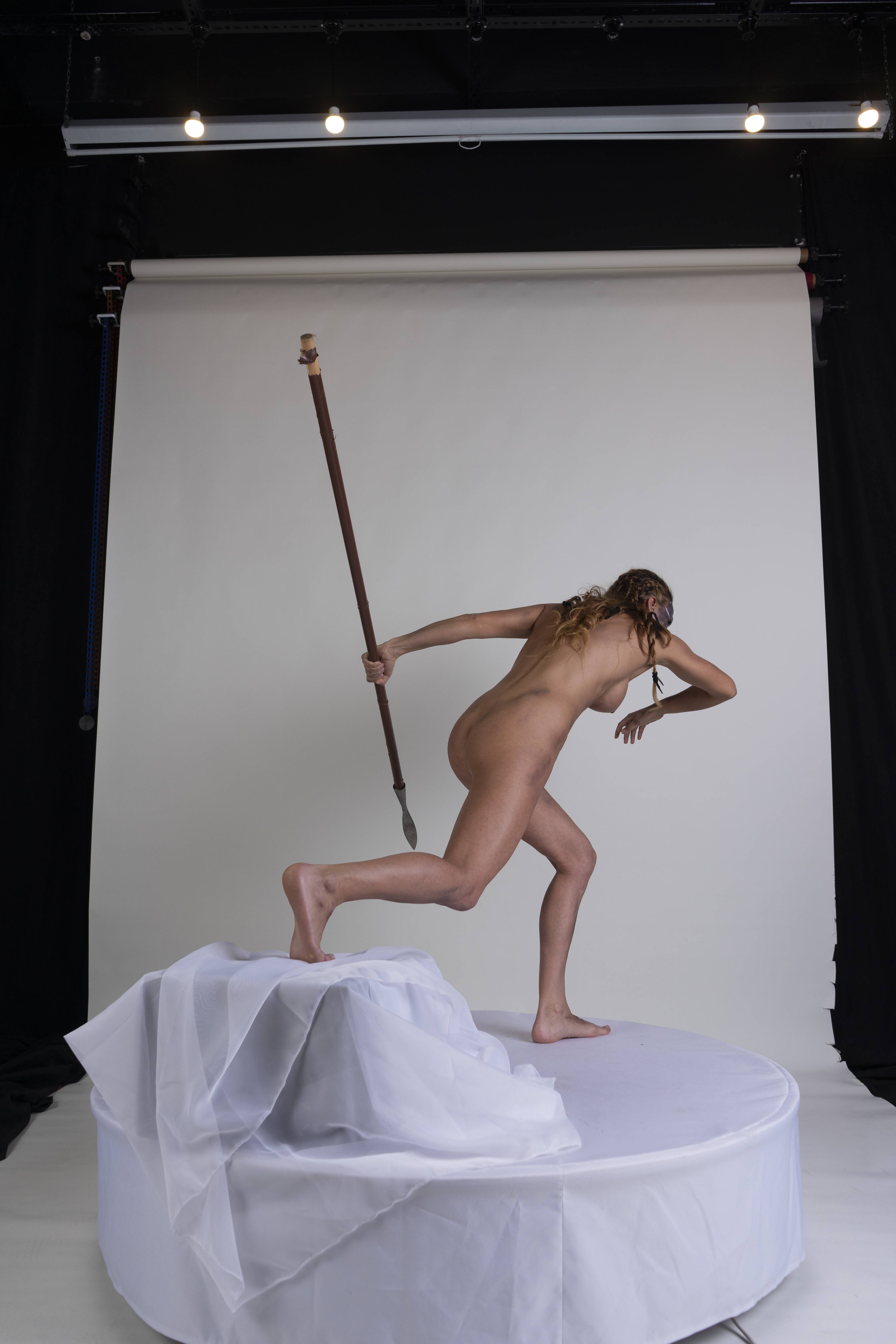 a nude woman stands on a white platform draped with a white cloth, holding a spear in a throwing position. she is set against a white backdrop that transitions to black on either side, suggesting a studio setting. the woman's body is toned and muscular. her stance is dynamic, as if in midaction. the overhead lighting indicates a controlled studio environment.