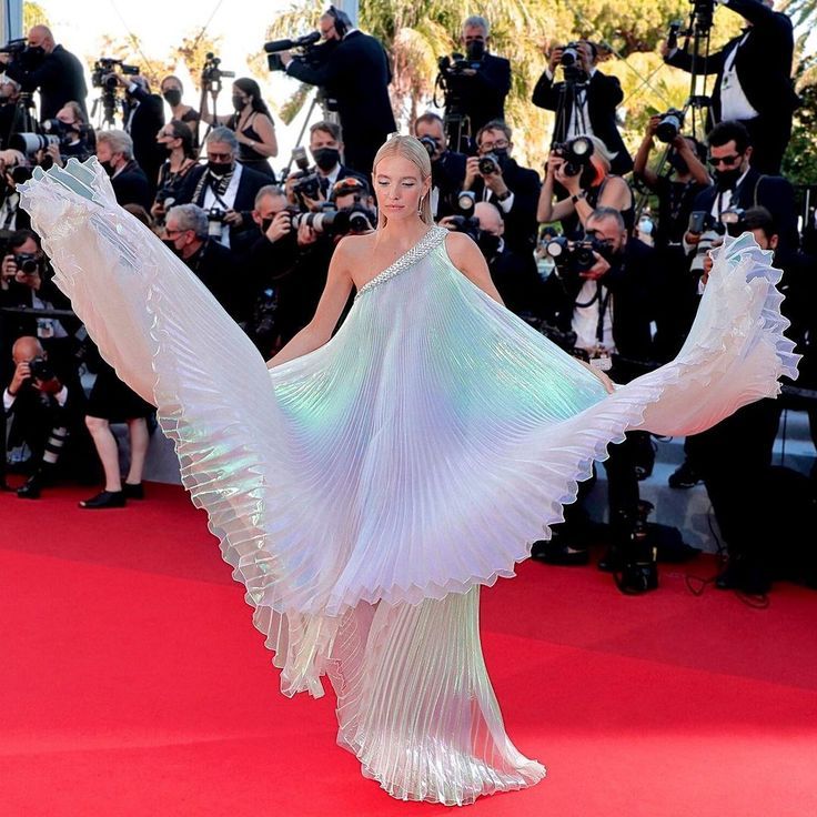 a woman in a white, iridescent, pleated gown stands on a red carpet, surrounded by photographers, as she poses for photos at an event, possibly the cannes film festival.  she is smiling and twirling her skirt, which flows beautifully in the breeze.