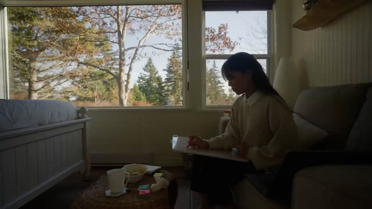 a young woman is sitting in a chair by a window, drawing on a tablet. the room is cozy and filled with natural light. outside the window, there are trees and a blue sky. the overall atmosphere is one of peace and relaxation.
