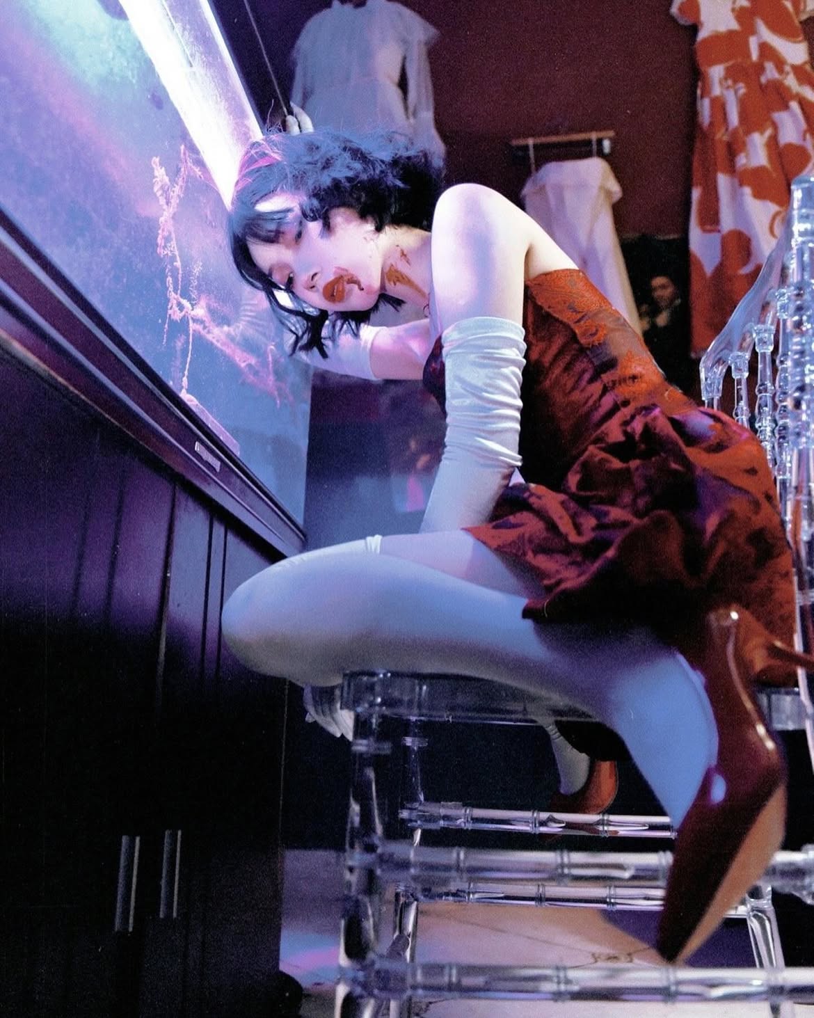 a woman in a red dress, white tights, and gloves, with bloodlike makeup, poses dramatically on a clear chair. she has pale skin and dark hair, suggesting a vampire or theatrical costume. the indoor scene features a dark cabinet, clothing racks with dresses, and a neon light, adding to the dramatic and theatrical atmosphere. the woman is posed artistically.