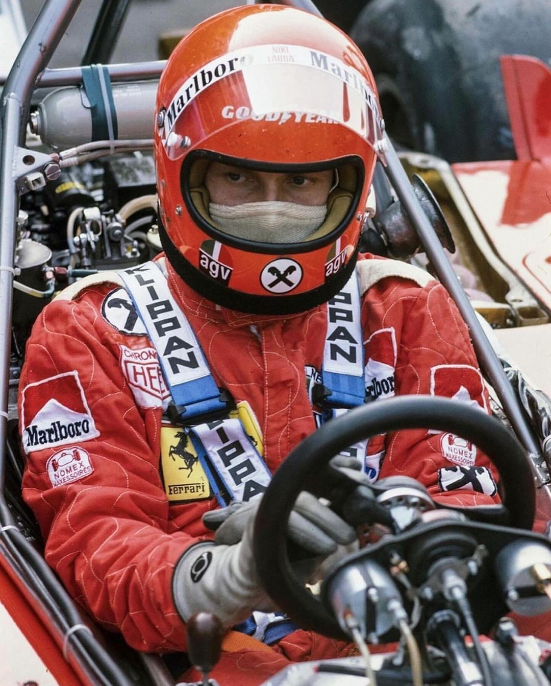 a closeup of a race car driver wearing a red helmet with marlboro and goodyear logos. the driver is wearing a red race suit with "marlboro" and "ferrari" patches. the driver is holding the steering wheel with his left hand, and his right hand is on the gear shift. the race car is in the background.