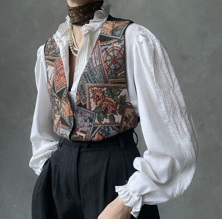 a person is shown from the waist up, wearing a white rufflecollared blouse, a tapestry vest, a pearl necklace, a scarf, and darkcolored pinstripe trousers. the background is a muted gray. the blouse has a ruffled collar and cuffs, and the vest is made of a tapestrylike fabric with various patterns and images. the pearl necklace is a multistrand piece, and the scarf is dark with a subtle pattern. the trousers are highwaisted and have a pinstripe pattern. the overall style is vintage and fashionable.