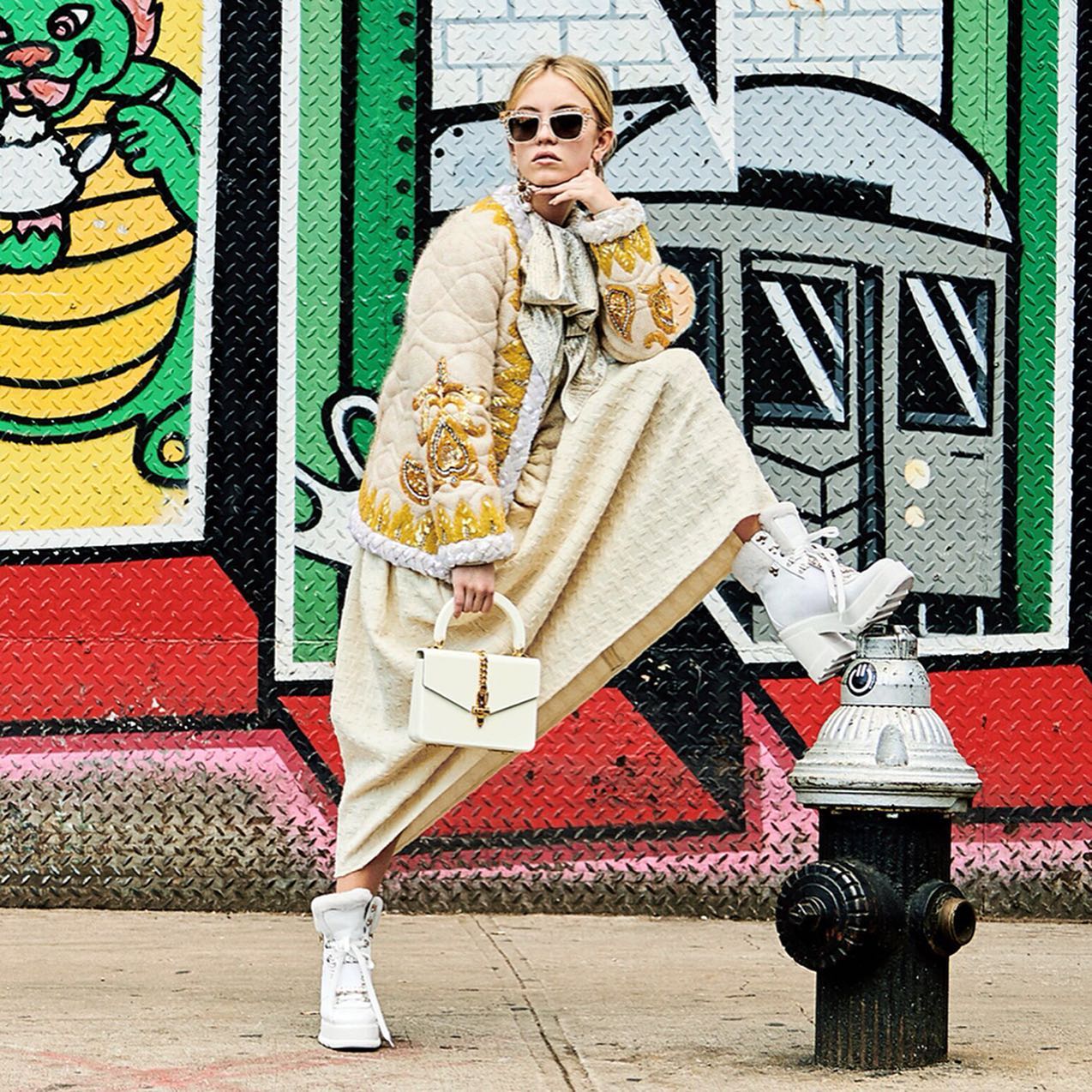the image showcases a young woman, possibly a model, stylishly dressed in a beige, embroidered quilted jacket, a long creamcolored skirt, and white platform hightop boots. she is carrying a white gucci handbag. she is posing playfully with one leg resting on a fire hydrant in front of a vibrantly colored graffiti wall in what appears to be an urban setting, possibly new york city. the overall style is contemporary and trendy, reflecting street fashion. the graffiti wall provides a bold backdrop contrasting with her elegant outfit.  the sunglasses and confident pose complete the stylish look.