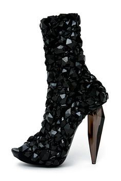 a black high heel boot with a pointy toe and a stiletto heel. the boot is made of a unique material that appears to be made of many small pieces of black material.