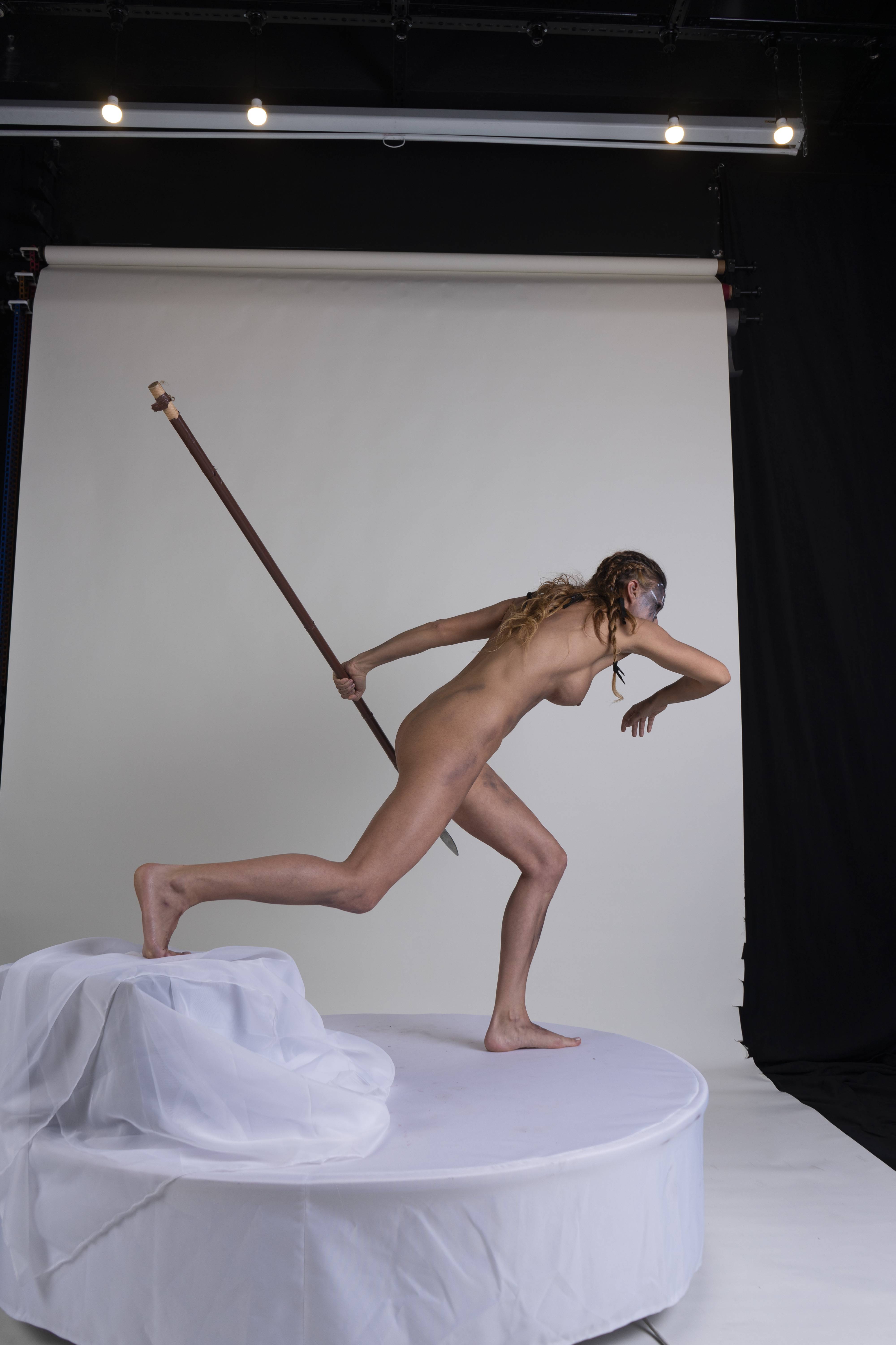 a nude woman with blue war paint on her face is running with a spear in a studio setting. she is posed on a round white platform covered with a white cloth. one foot is placed on the clothcovered section of the platform, as if she has just stepped on it. her right arm is extended forward, and she holds a spear in her left hand, angled down and back. the background is a white backdrop, and the scene is lit by studio lights overhead.