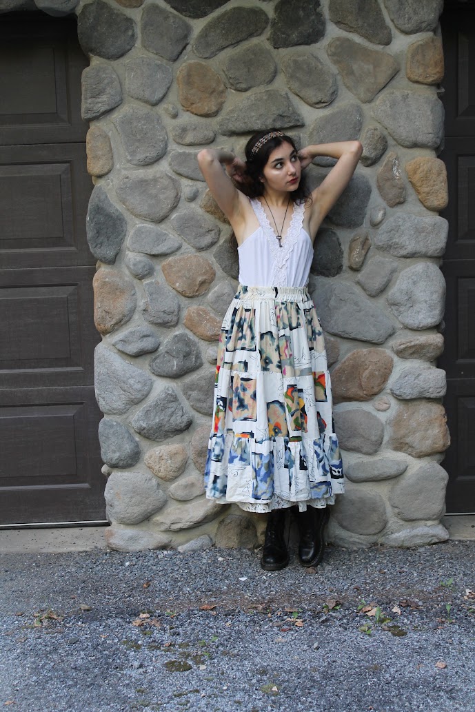 a fulllength shot of a young woman standing against a stone wall. she is wearing a white cropped top and a long, colorful, patterned skirt. she is also wearing black boots and a headband. the woman is posing with her arms behind her head, and she appears to be looking off to her left side. the image has a casual, summery feel to it.