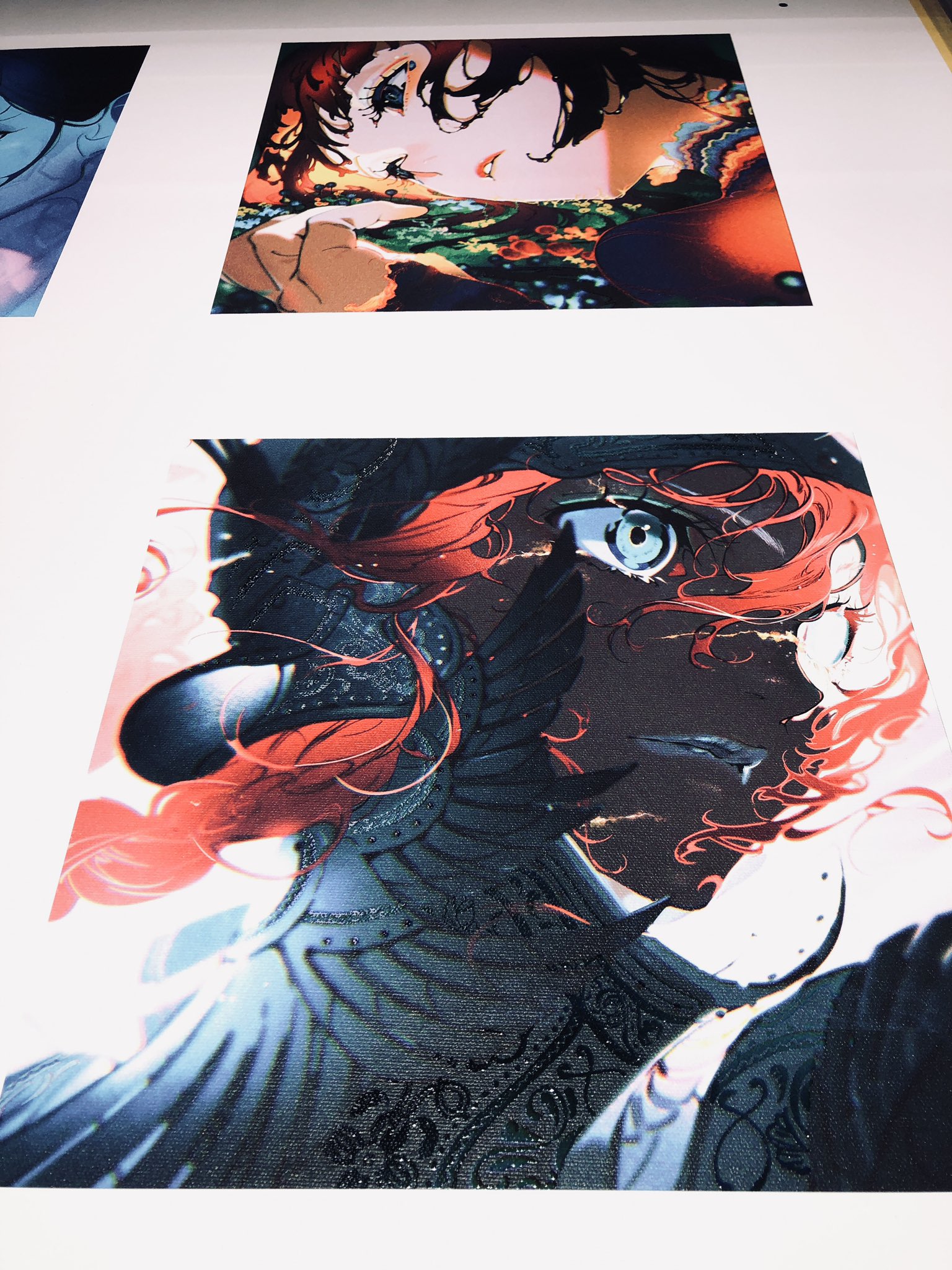 the image shows two closeup portraits of a woman with vibrant red hair and striking blue eyes, seemingly printed on canvas and displayed on a wall. the illustrations showcase a style reminiscent of manga or anime, with detailed linework and vibrant colors. the top portrait captures the subject from the shoulders up, her head slightly tilted and gazing downwards, while the bottom one presents a more dramatic, angled perspective, focusing on her face and the intricate details of her attire.  the overall aesthetic exudes a sense of fantasy and strong character design.