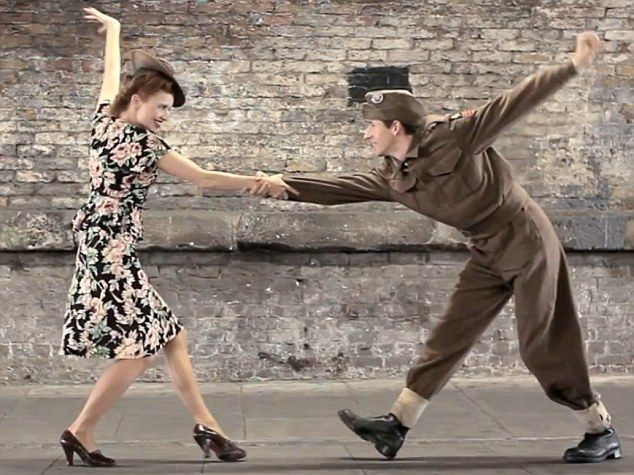 a woman in a floral dress dances with a man in a military uniform. they are both smiling and enjoying themselves. the background is a brick wall.