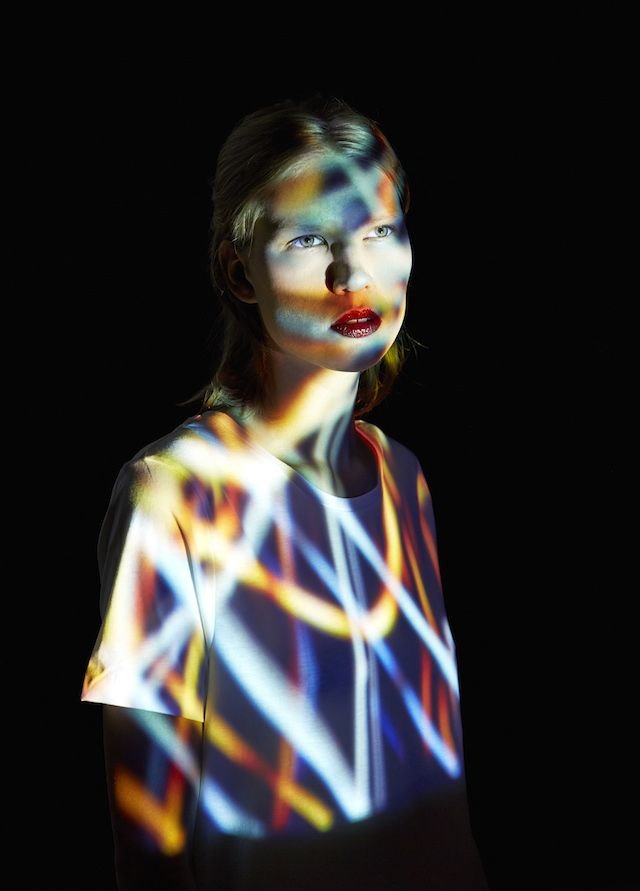 a woman with blonde hair and red lipstick poses against a dark background. she is wearing a white tshirt and her face is illuminated by a colorful projection that creates an abstract and artistic effect. the colors are bright and vibrant, and the overall mood of the image is mysterious and seductive.