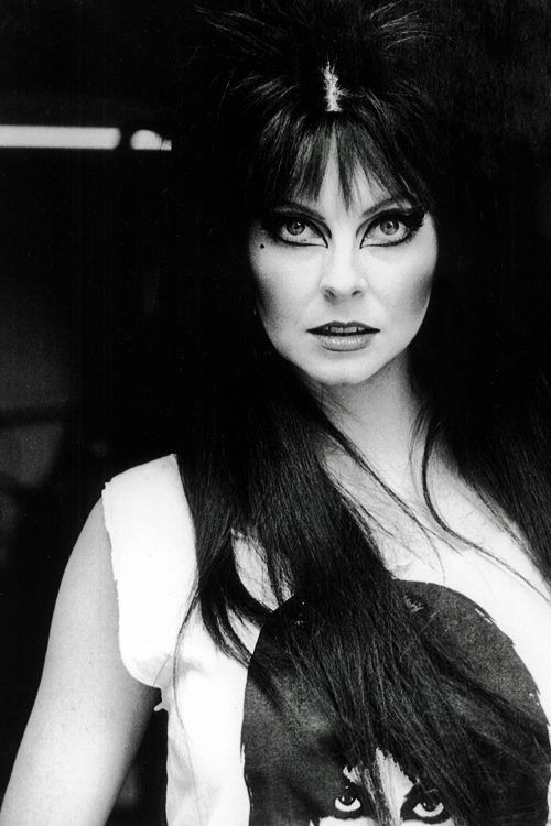 a black and white portrait of a woman with long black hair. she is wearing dark makeup and dark lipstick. she is wearing a white tshirt. the photo was taken in the 1970s.