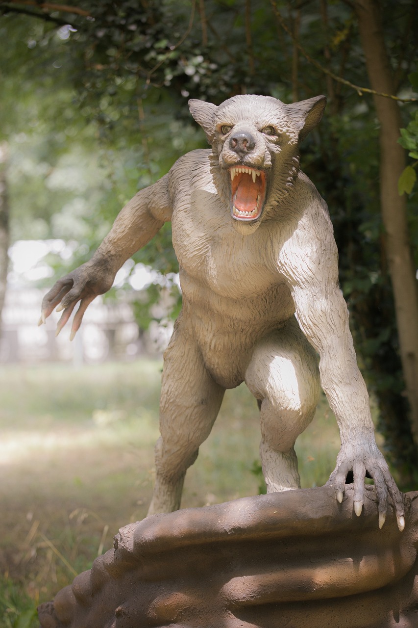 a werewolf sculpture stands in a forest setting. the creature has a doglike head with sharp fangs visible in its open mouth. it is bipedal, with humanlike arms and hands ending in long, sharp claws. the sculpture is made of a grey material and is positioned on a rocky base. the background includes trees and foliage, suggesting a natural environment.
