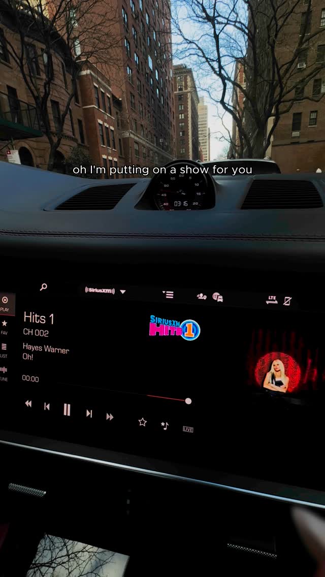 the image shows the dashboard of a car with the infotainment system displaying siriusxm radio, playing the song "oh!" by hayes warner. the view through the windshield reveals a city street with tall buildings and trees. text overlay reads "oh i'm putting on a show for you."