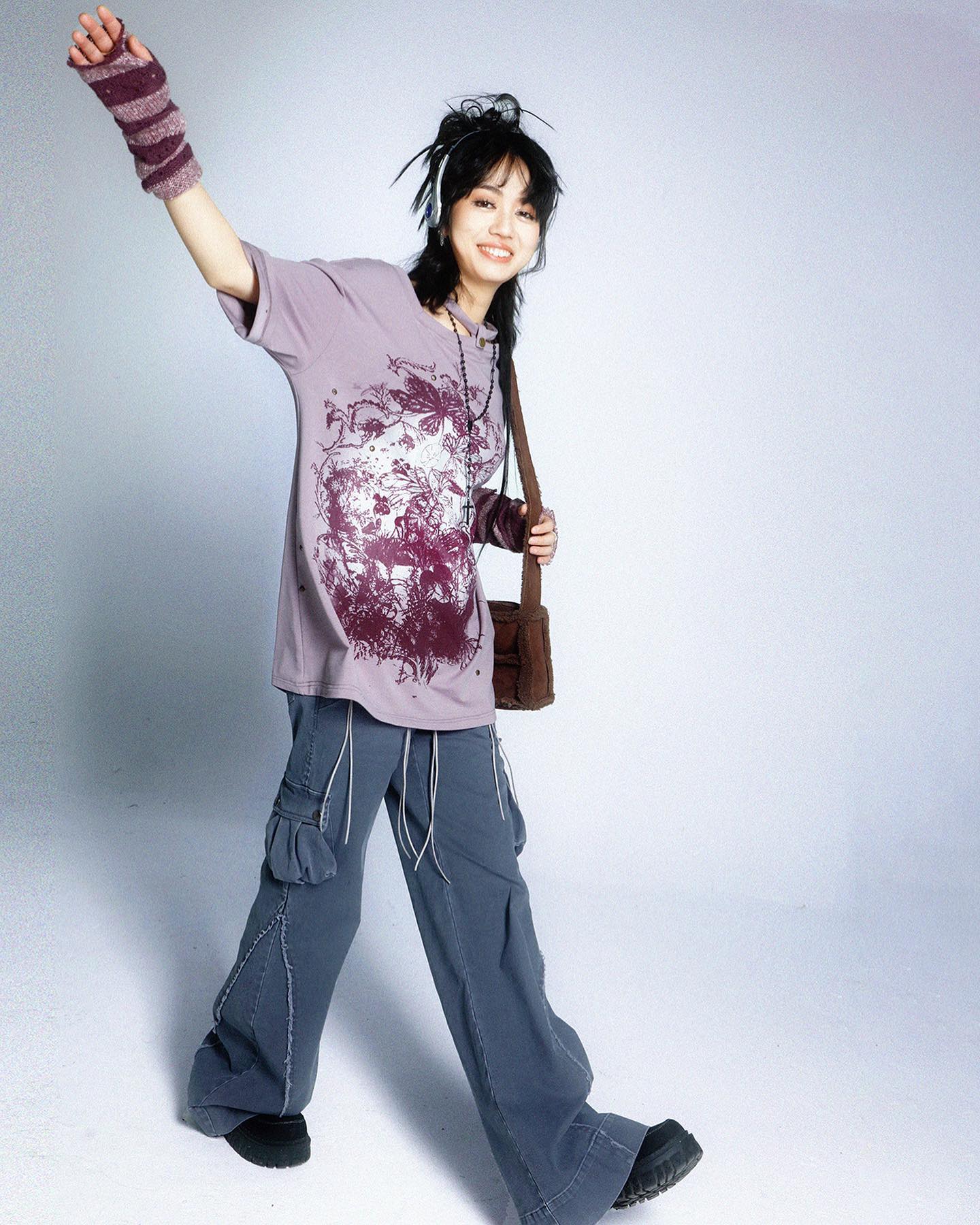the image shows a young woman posing against a light grey backdrop. she's wearing a loosefitting, lavendercolored graphic tshirt with a dark purplish print, paired with widelegged, grey cargo pants.  she also sports fingerless gloves, a brown shoulder bag, and what appears to be headphones around her neck. her dark hair is styled casually. the overall style blends casual elements with a hint of grunge or 90s/y2k aesthetics. the woman is smiling and positioned dynamically, with one arm raised. the setting is simple and focuses on the clothing and the model's pose.