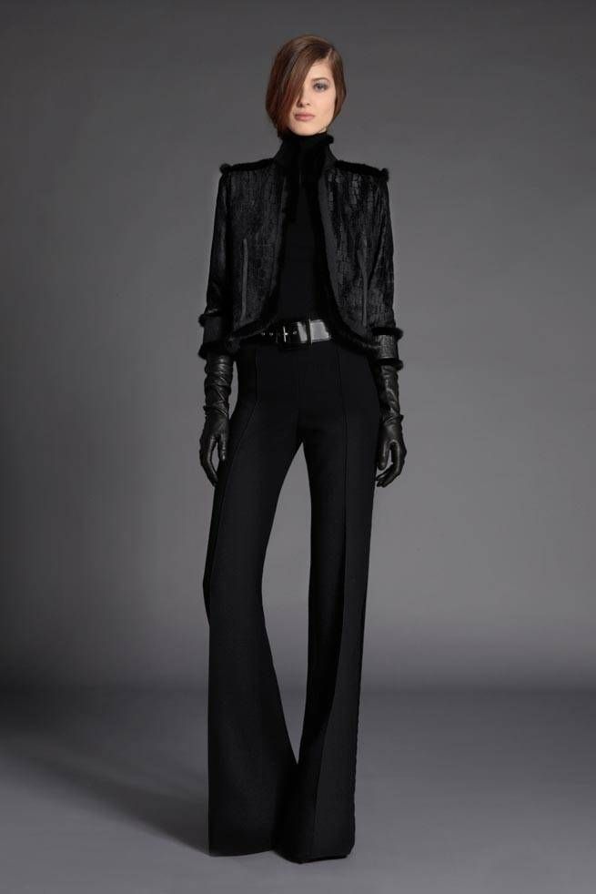 a young woman in a black furtrimmed jacket, black flared trousers, a black turtleneck sweater, and black leather gloves stands against a gray background. she is looking at the camera, with a slightly serious expression on her face. her outfit is elegant and stylish, and she appears to be confident and poised.