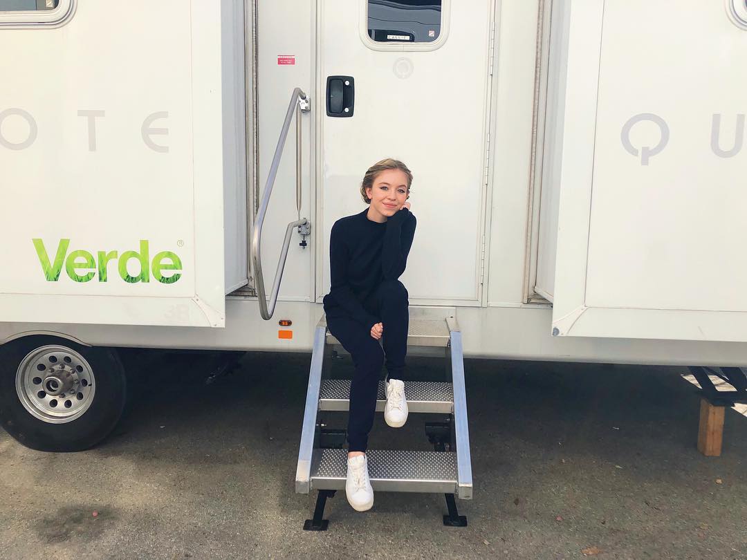 a young woman, possibly an actress, is sitting on a set of metal steps in front of a white production trailer. the trailer has the word "verde" and a logo on it. the woman is wearing a black sweater and pants with white sneakers and is smiling, looking comfortable and relaxed. the setting appears to be a film or movie set.