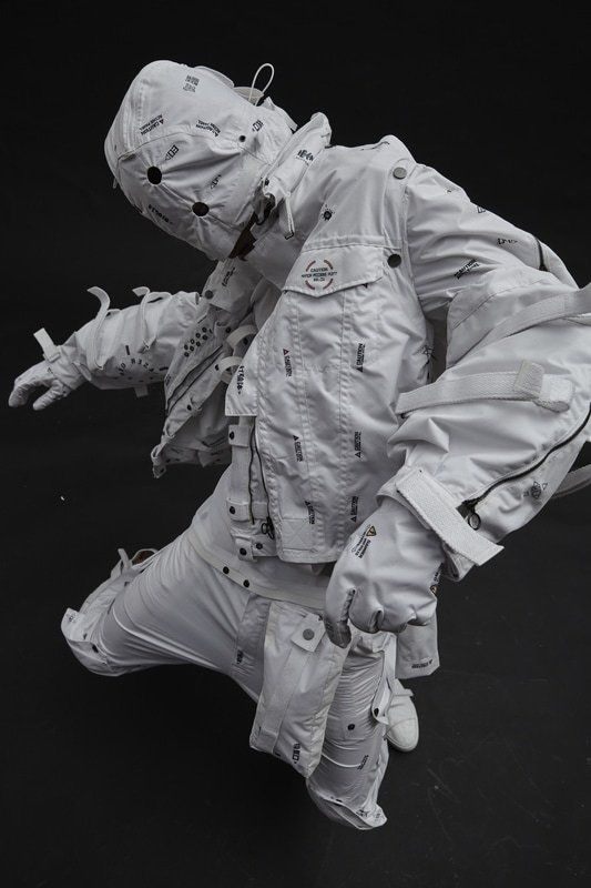 a model in all white futuristic clothing, posed with one leg raised, on a black backdrop. the jacket, pants, and boots are all white, and the model is wearing a white helmet. the clothing is heavily detailed with straps, buckles, and other hardware. the pose is dynamic and edgy, and the overall look is very contemporary and cool. the black and white aesthetic is sleek and modern, and the clothing is both stylish and functional. the model's pose and the clothing's intricate details make this a striking image.