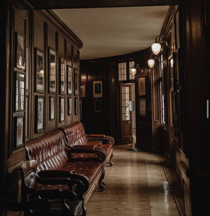 the image showcases a long, dimly lit hallway or corridor in a grand, old house or mansion. the walls are adorned with dark, rich wood paneling, and a gallery wall displays numerous framed portraits and artworks.  the hallway is furnished with luxurious, deep brown leather sofas and armchairs, all with tufted upholstery. warm lighting from wall sconces casts a soft glow on the scene, highlighting the ornate details of the architecture and furniture. the overall ambiance is elegant, sophisticated, and subtly mysterious, hinting at a historic or heritage setting. the wooden floor adds to the classic and opulent feel.