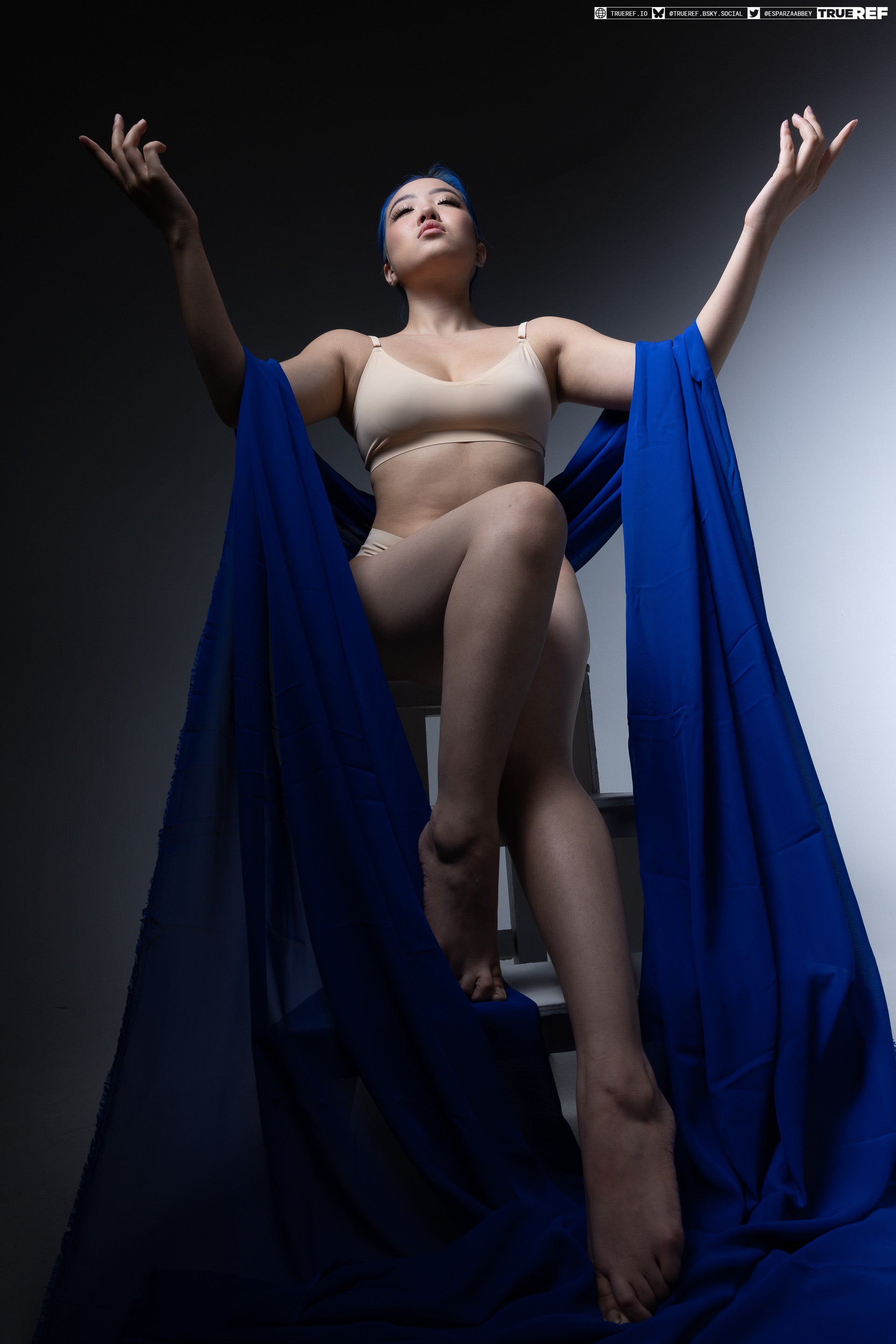 a studio shot of a woman with blue hair wearing a bra and underwear, seated on a ladder with blue fabric draped around her. she has her arms raised and is looking up. the photo is taken from a low angle with studio lighting.