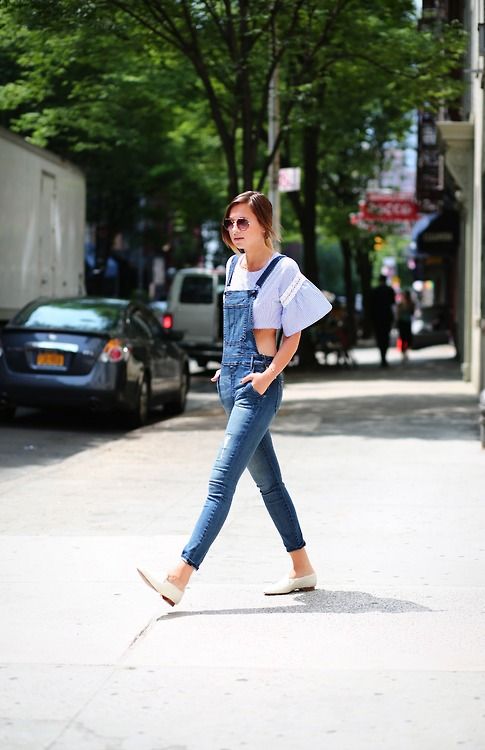 a stylish woman is walking down a city street wearing denim overalls over a light blue cropped top with bell sleeves.  she's wearing white flats and sunglasses. the setting is sunny and outdoors. the overall style is casual yet trendy.
