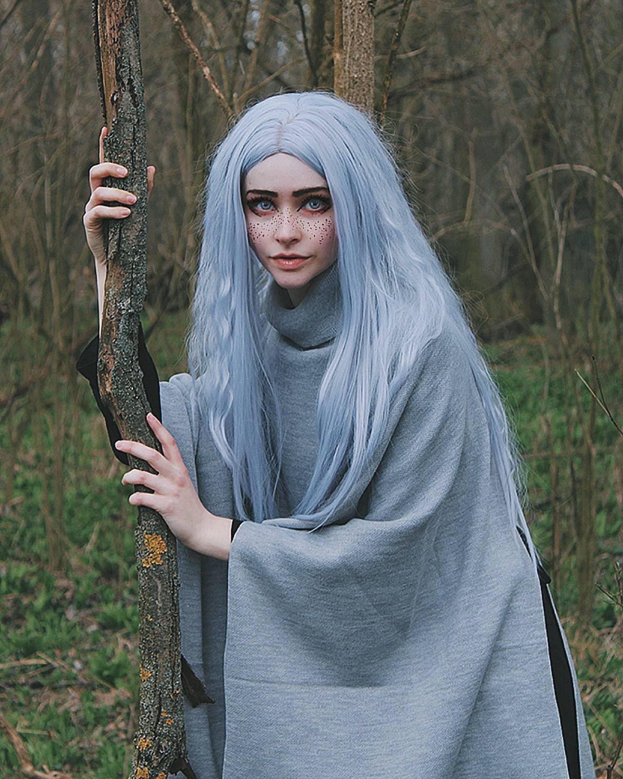 a captivating portrait of a young woman with long, light blue hair, styled in an elf or witchlike costume. she's dressed in a flowing gray cape or poncho and stands in a forest setting, holding a wooden branch or staff. the overall mood is mystical and ethereal, enhanced by the soft lighting and natural surroundings.  her pale skin and subtle freckles add to the otherworldly aesthetic. the image suggests a fantasy or cosplay theme.